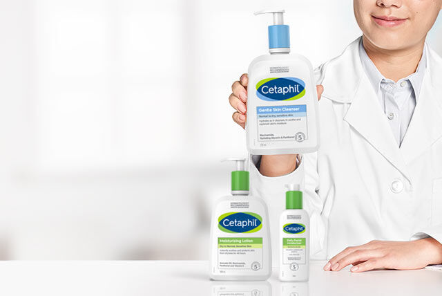 Person i hvit frakk holder en flaske Cetaphil Gentle Skin Cleanser, med andre Cetaphil-produkter på et nærliggende bord