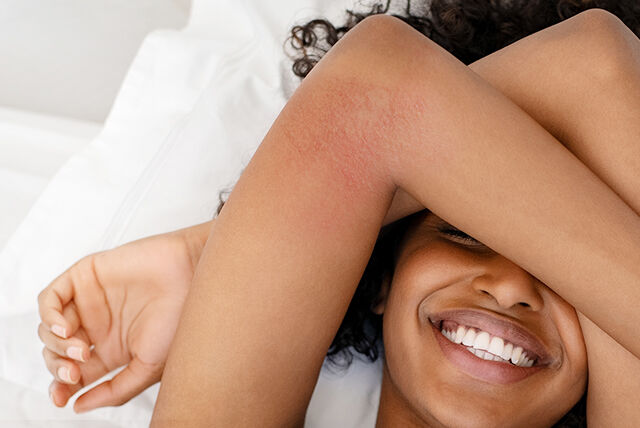 En smilende kvinne ligger på ryggen i sengen, med armene krysset over øynene