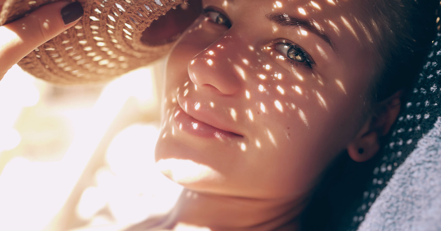 En smilende kvinne ser inn i kameraet, ansiktet i skyggen av stråhatten, bortsett fra noen stråler av lys som har kommet gjennom hullene i hatten